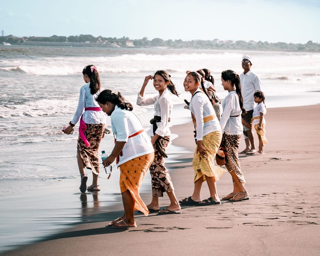 People in traditional Balinese dress walking along a black-sand beach during a ceremony